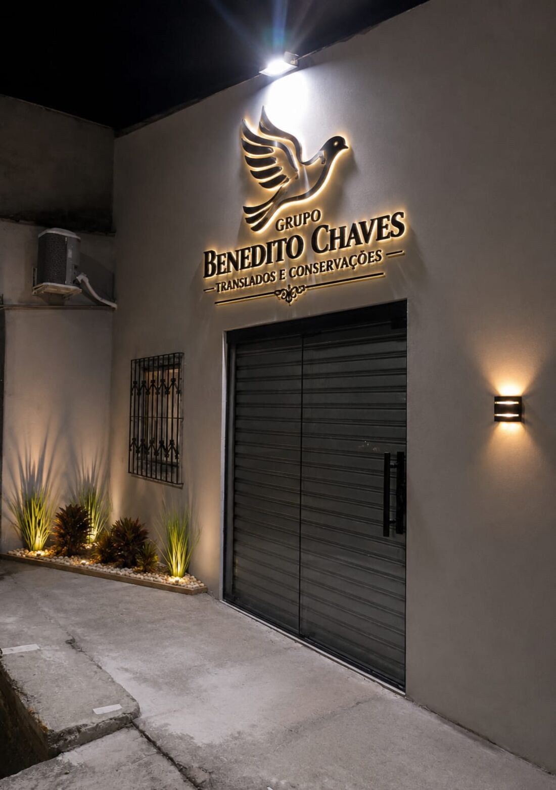 Entrada da sede do Grupo Benedito Chaves à noite, com a pomba da marca em letreiro iluminado.