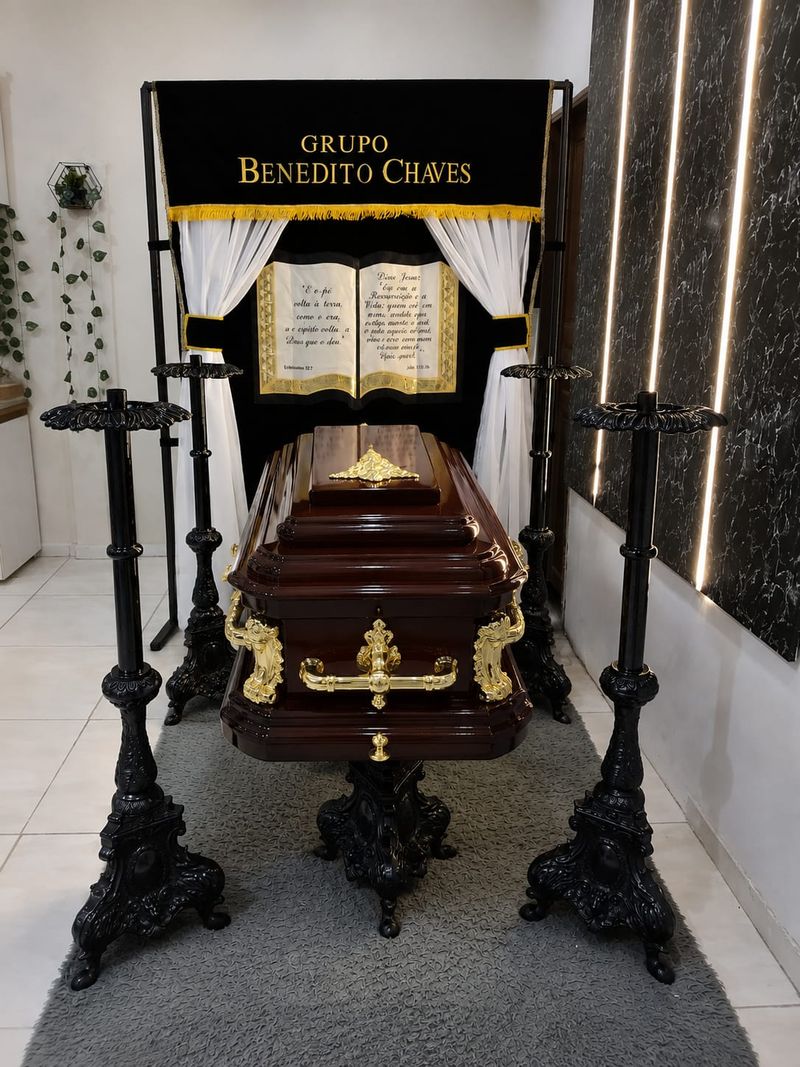 Esquife em madeira mogno com alças e ornamentos dourados, montado na capela do Grupo Benedito Chaves entre castiçais, sob o pálio bordado com o nome da empresa.